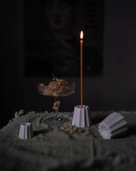 Candle holder in the shape of a traditional French dessert canelé in a matte white finish on a table with moss green table cloth. Candle is lit and surrounding environment is dark and dimmed. 