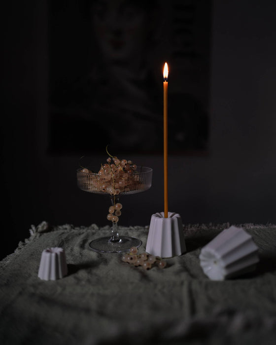 Candle holder in the shape of a traditional French dessert canelé in a matte white finish on a table with moss green table cloth. Candle is lit and surrounding environment is dark and dimmed.