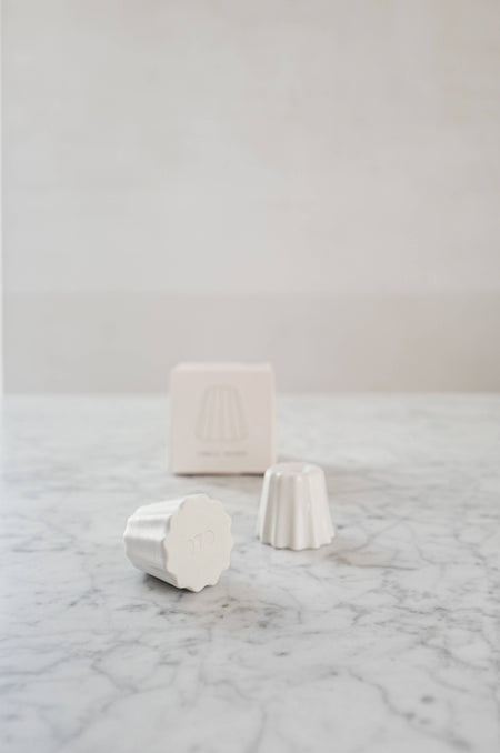 Two Small candle holder in the shape of a traditional French dessert canelé in a matte white finish. showcased on a marble table with one packaging.
