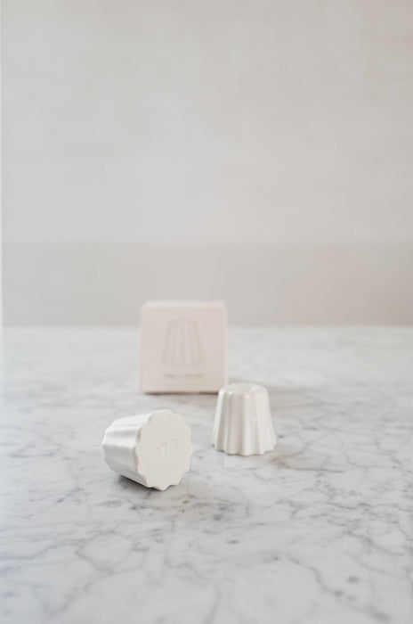 Two Small candle holder in the shape of a traditional French dessert canelé in a matte white finish. showcased on a marble table with one packaging.