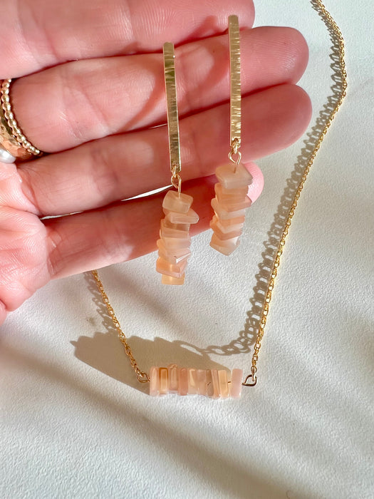 A set of Peach Moonstone dangle earrings. Ten smallm squared, peach moonstone squares hanging off a 14K gold-filled one inch long bar. Earrings held in hand. Below is a 14K gold-filled necklace with ten Peach Moonstone square charms, side by side stacked on a bar.