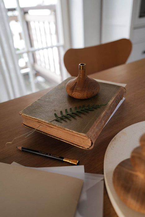 Oiled oak candle stick holder with brass detail displayed in a home setting. Candle holder placed on a brown book which is placed on a wooden table. 