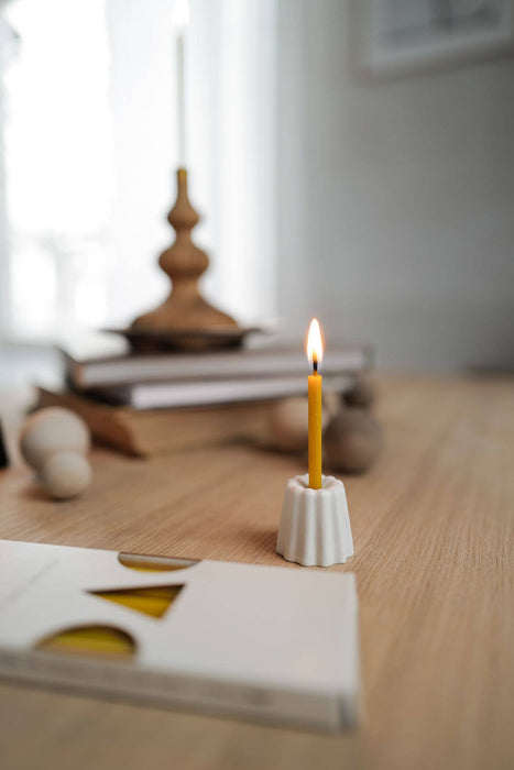 Small candle holder in the shape of a traditional French dessert canelé in a matte white finish with a lit slim 100% beeswax candle displayed on a wooden table in a home setting.