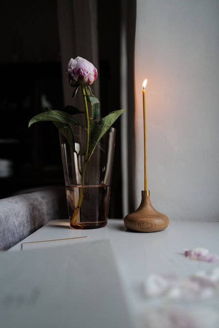 Oiled oak candle stick holder with brass detail and slim beeswax candle in a home setting displayed next to a small vase with flower.