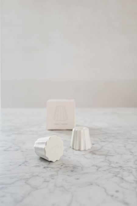 Two Small candle holder in the shape of a traditional French dessert canelé in a matte white finish. showcased on a marble table with one packaging. 