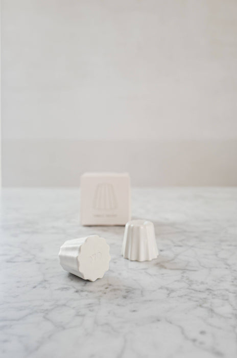 Two Small candle holder in the shape of a traditional French dessert canelé in a matte white finish. showcased on a marble table with one packaging. 