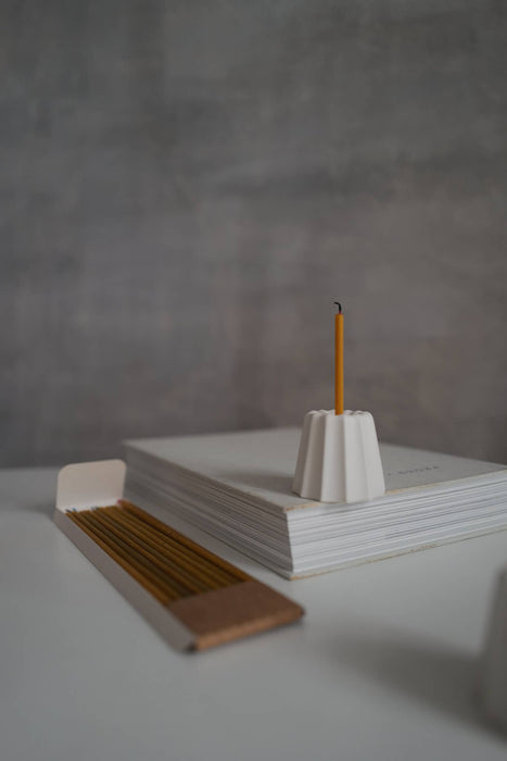 Candle holder in the shape of a traditional French dessert canelé in a matte white finish showcased in a home setting on a pile of papers and next to a package of 100% beeswax candles.