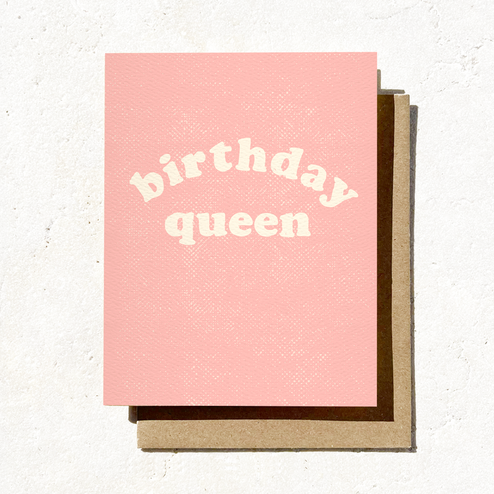 A pink A2 size greeting card with 'birthday queen' printed in bold letters, accompanied by a brown envelope, against a white textured background.