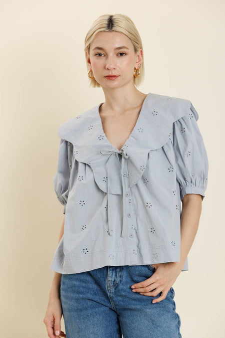 Woman wearing a light blue blouse with ruffled sleeves and a bow tie, standing against a beige background.