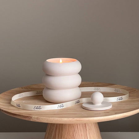 A donut-shaped soy wax candle with a wood wick, placed on a wooden stand with the brand name 'estelle' visible on the candle and the stand.
