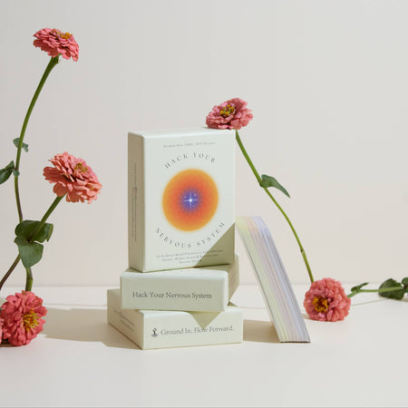 A card deck titled 'Hack Your Nervous System' displayed alongside a set of three cards, with beautiful pink flowers arranged in the background.