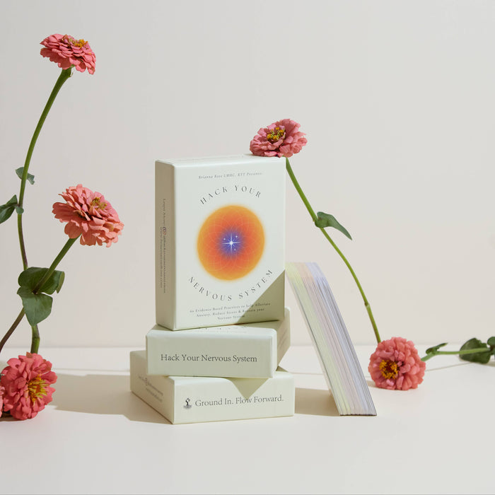 A card deck titled 'Hack Your Nervous System' displayed alongside a set of three cards, with beautiful pink flowers arranged in the background.