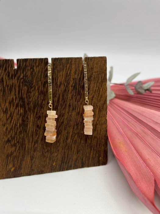 A set of Peach Moonstone dangle earrings. Ten small squared, peach moonstone squares hanging off a 14K gold-filled one inch long bar. Earrings are hanging off a wooden display.