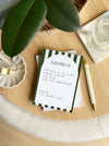 Notebook with green and white striped cover on a wooden surface with a pen and plant leaves.