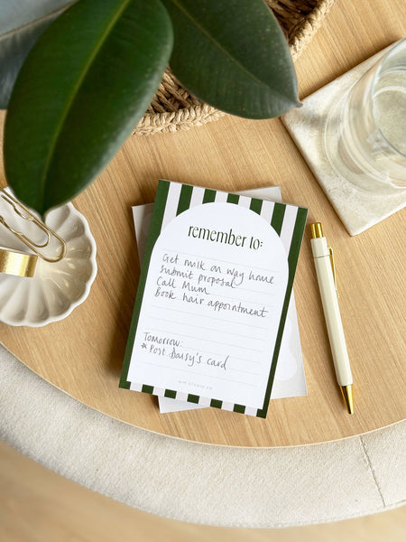 Notebook with green and white striped cover on a wooden surface with a pen and plant leaves.