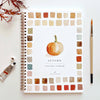 Watercolor workbook titled 'Autumn' with color swatches and a pumpkin illustration, surrounded by art supplies on a white background.