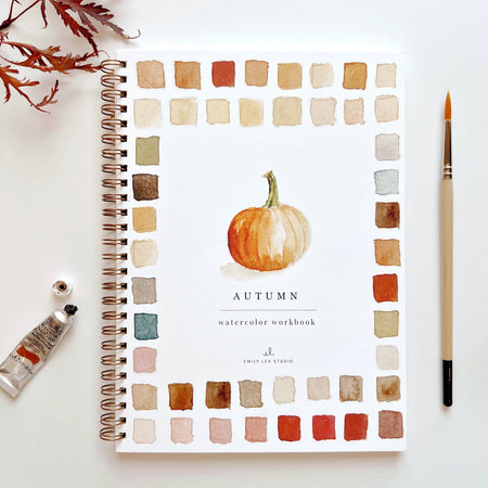Watercolor workbook titled 'Autumn' with color swatches and a pumpkin illustration, surrounded by art supplies on a white background.