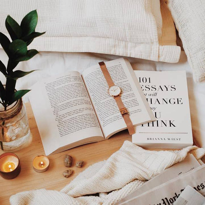 Open book with a watch on top, next to a candle and plant on a wooden surface.